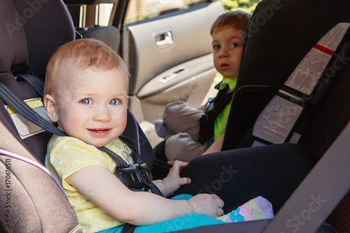 Portrait of two cute white Caucasian toddlers childen sitting in car seats looking in camera. Smiling babies in automobile vehicle fastened with seatbelts.