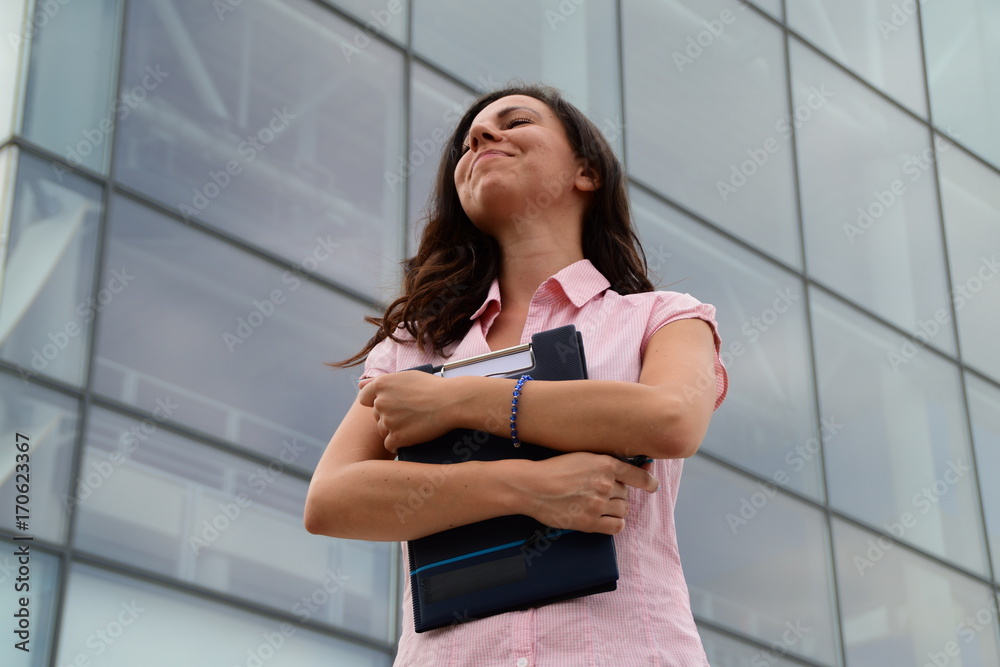 Smiling and proud, young business woman holding notes in her hands, standing in front of a glass building Smiling and proud, young business woman holding notes in her hands, standing in front of a glass building