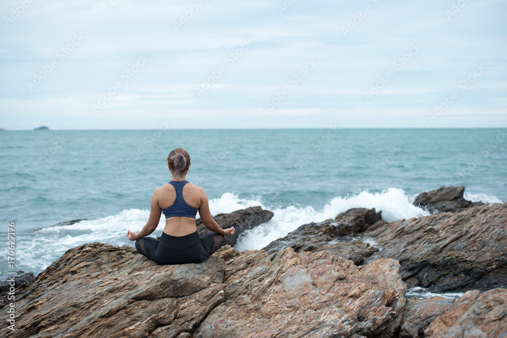 Young asian woman yoga and meditation on the stone at the sea beach, healthy lifestyle and fitness concept.