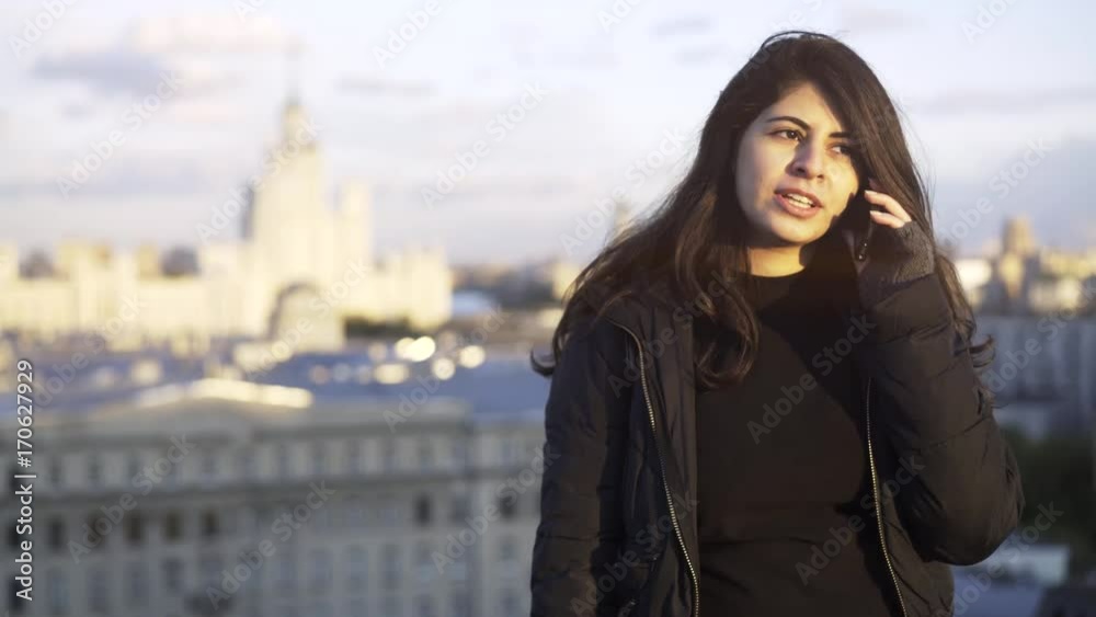 Close up of a young woman with long black hair wearing a black vest and talking on a phone while standing on a roof of a building in a city. Locked down real time close up shot Close up of a young woman with long black hair wearing a black vest and talking on a phone while standing on a roof of a building in a city. Locked down real time close up shot