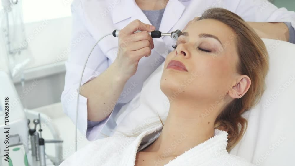 Female cosmetologist moving iontophoresis roller along client's forehead. Professional beautician performing galvanic facial treatment. Pretty blond woman getting anti-aging procedures for her skin Female cosmetologist moving iontophoresis roller along client's forehead. Professional beautician performing galvanic facial treatment. Pretty blond woman getting anti-aging procedures for her skin