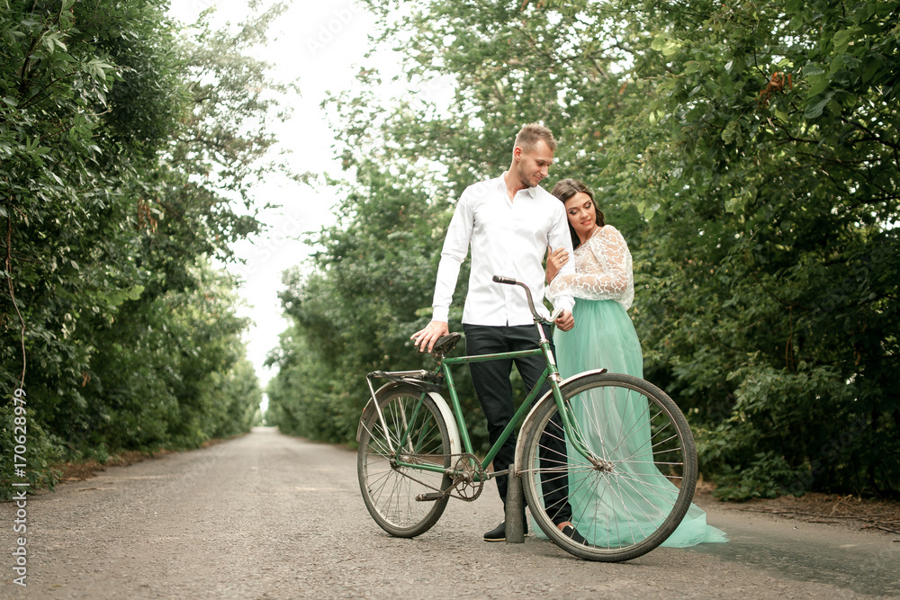 Bride and groom stand next to bicycle on forest road. Bride and groom stand next to bicycle on forest road.