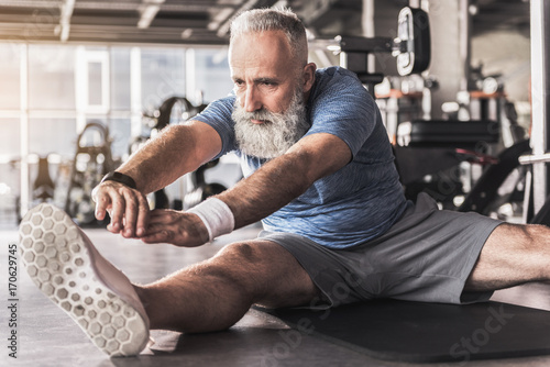 Serious old bearded man is enjoying exercises in fitness center