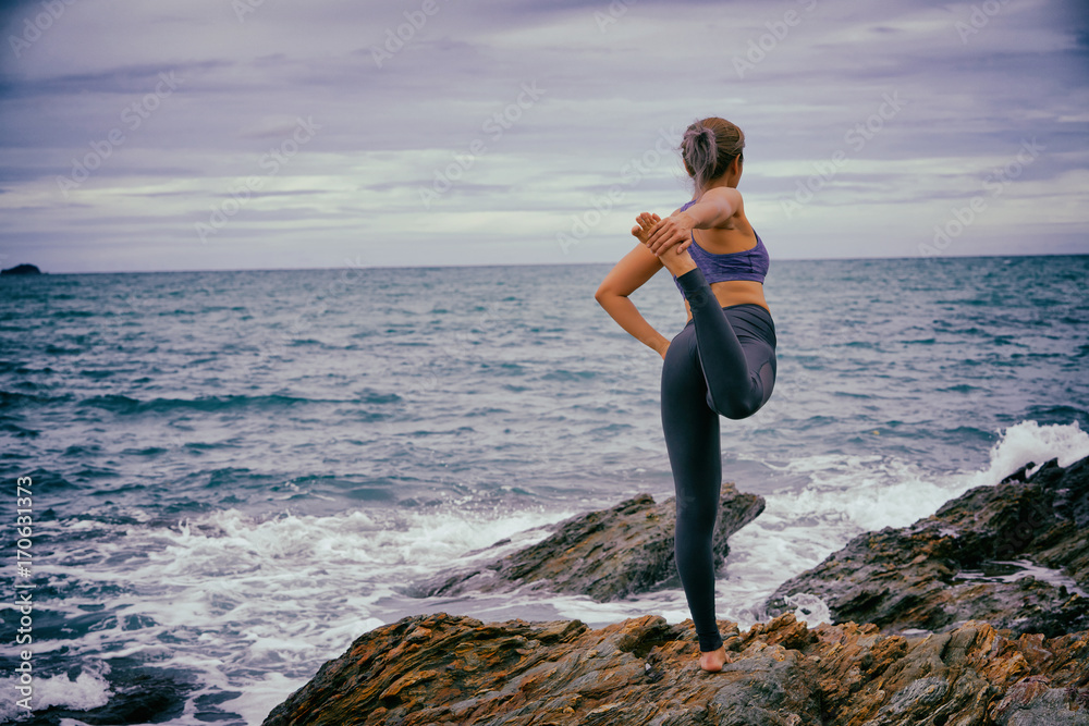 Asian woman standing yoga postures Standing Bow Pulling Pose on the rocks. Sea and sky as background.