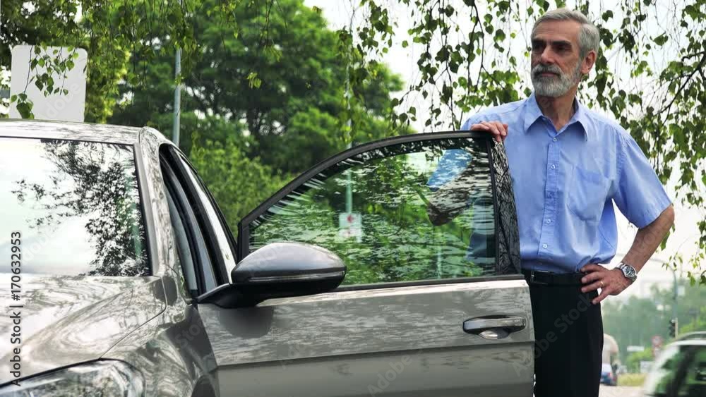 Senior man stands next to the car and looks around (car stands on the verge of road) - street in the background Senior man stands next to the car and looks around (car stands on the verge of road) - street in the background