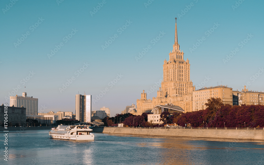 Obraz premium Autumn cityscape on sunny evening: Moscow river with touristic ship on it, historical hotel building calling 