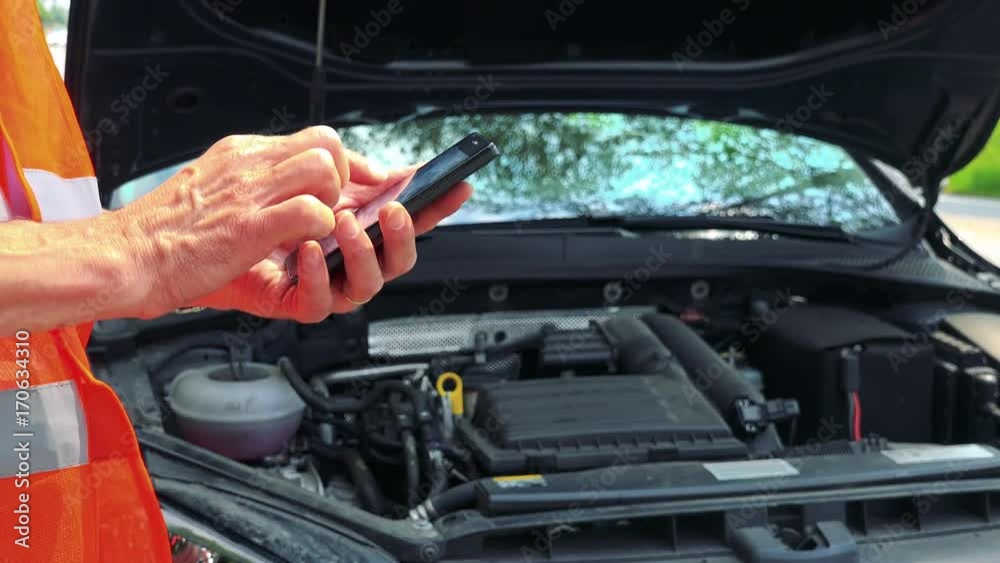 Senior man wears warning vest and works on smartphone next to the open hood broken car - closeup Senior man wears warning vest and works on smartphone next to the open hood broken car - closeup