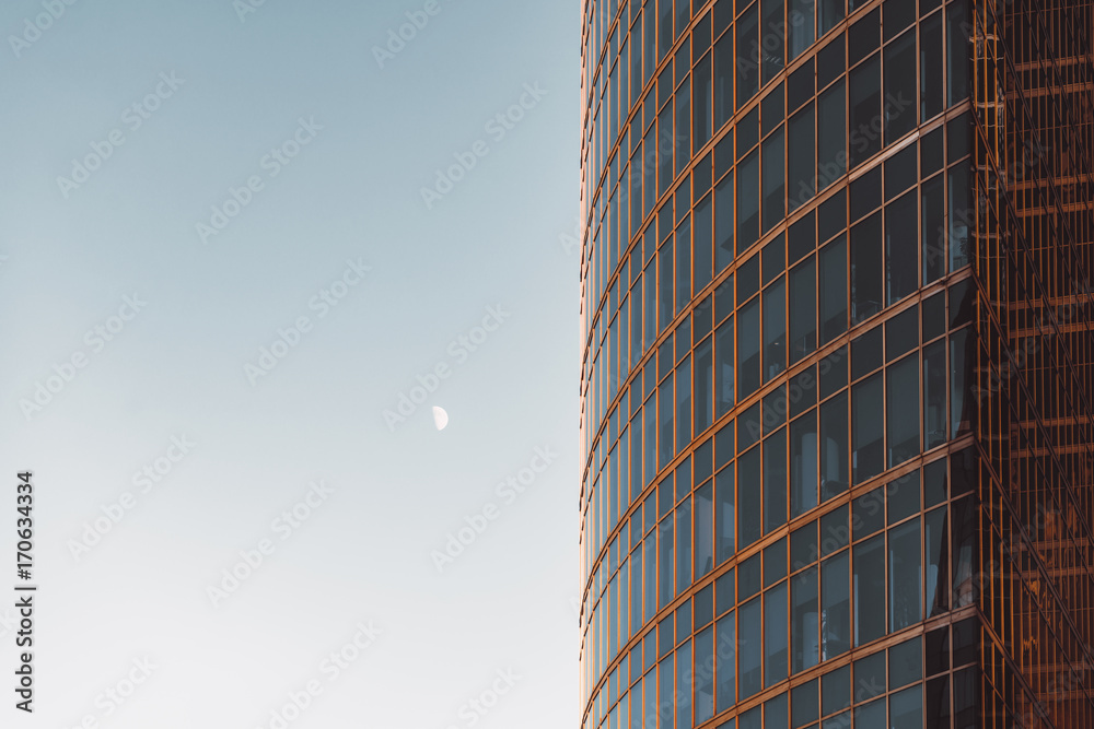 Obraz premium Side view of modern skyscraper teal and orange glassy facade with blue sky and moon on other side, offices overlooked behind windows,