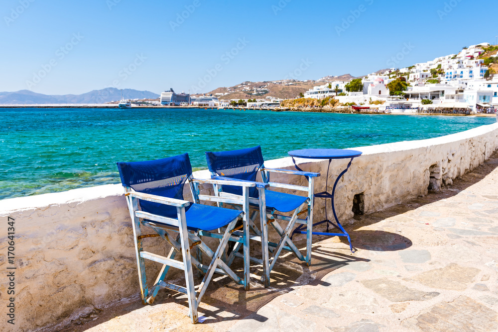 Blue chairs and table on the coastal promenade with beautiful view of the sea and port in Mykonos town. Cyclades Islands, Greece.