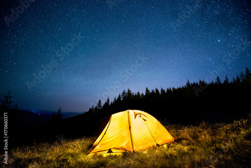 Glowing camping tent in the mountain forest under a starry sky