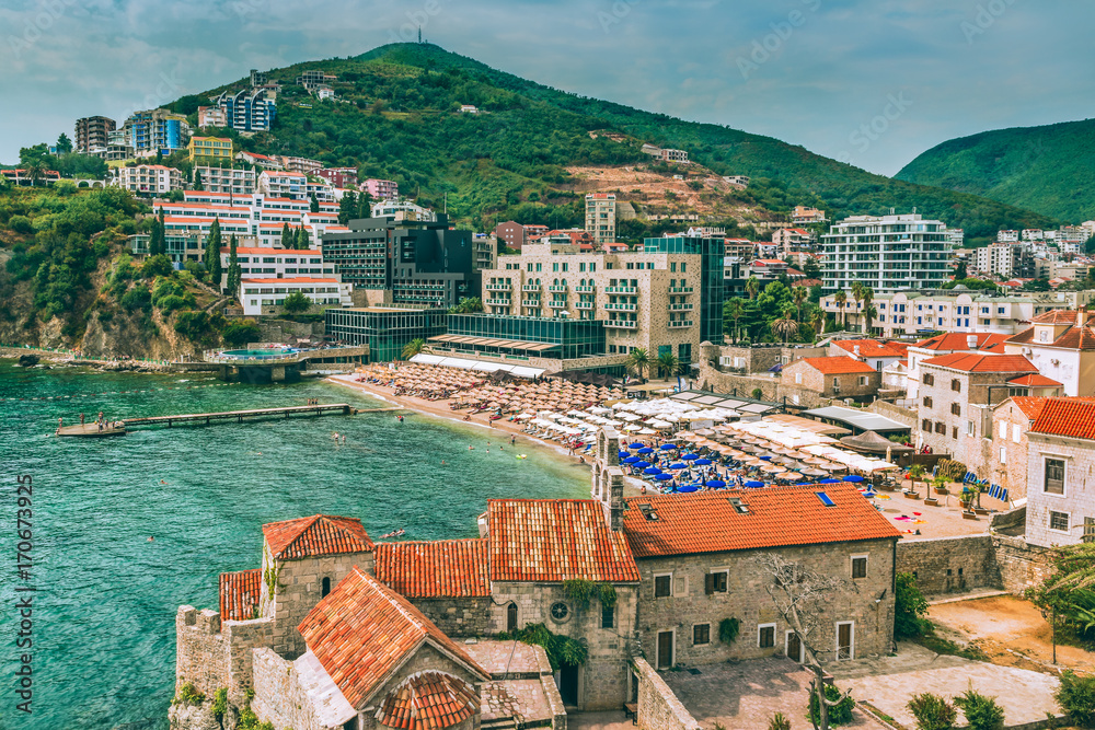 Fragment of the old town of Budva: ancient walls and a red tiled roof and a view of a modern beach, Montenegro, Europe. Budva is one of the best and most popular resorts of the Adriatic Riviera.