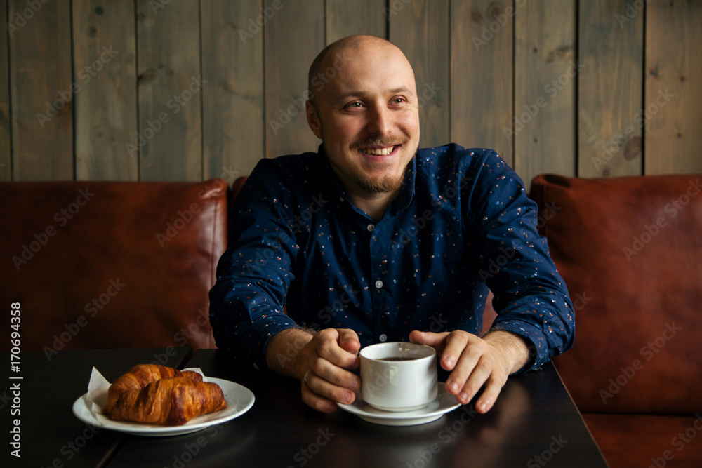 Young serious fashionable man sitting alone in loft-styled cafe and smiling. Cup of coffee and croissants on the table. Former factory building, natural daylight. Young serious fashionable man sitting alone in loft-styled cafe and smiling. Cup of coffee and croissants on the table. Former factory building, natural daylight.