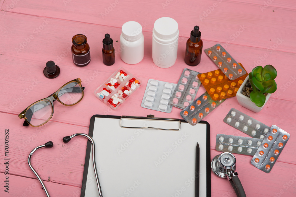 Φωτογραφία Doctor workplace with blank clipboard, medicine bottles, red stethoscope, eyeglasses and pills on pink wooden table
