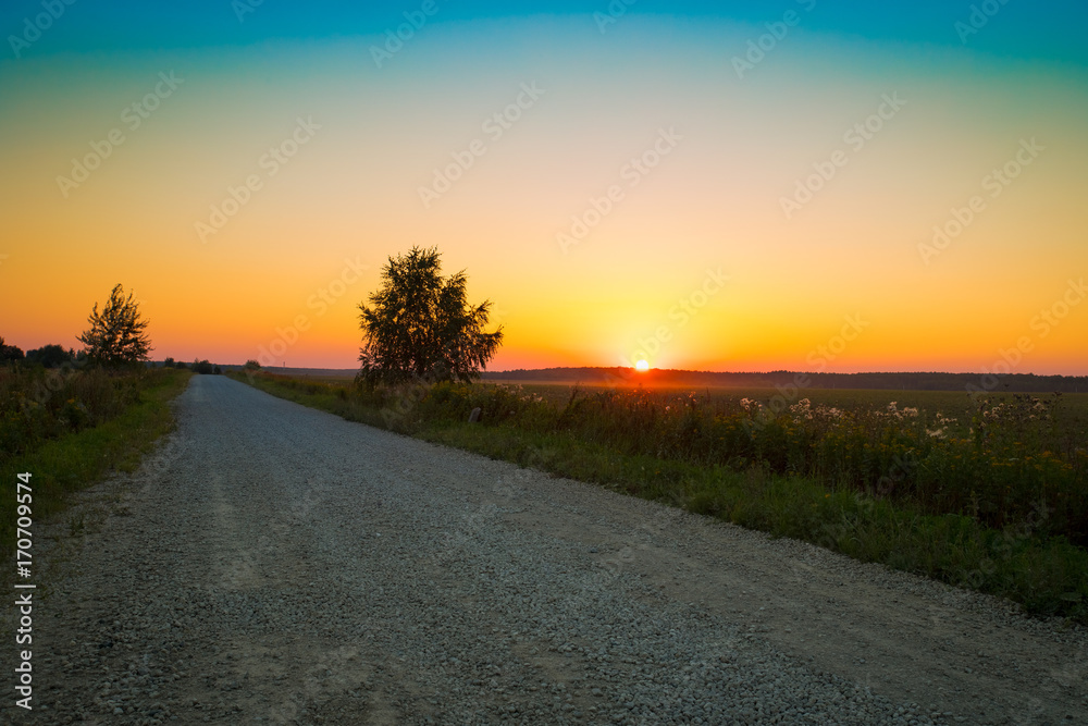 Beautiful Summer Landscape With Rural Countryside Road Covered by Rubble Under Bright Colorful Of Yellow, Orange, Blue Colors Sunset Or Sunrise In Evening Day.