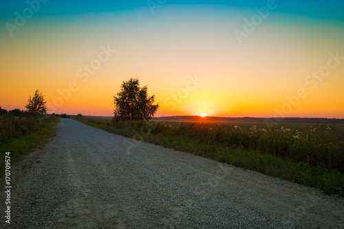 Beautiful Summer Landscape With Rural Countryside Road Covered by Rubble Under Bright Colorful Of Yellow, Orange, Blue Colors Sunset Or Sunrise In Evening Day.
