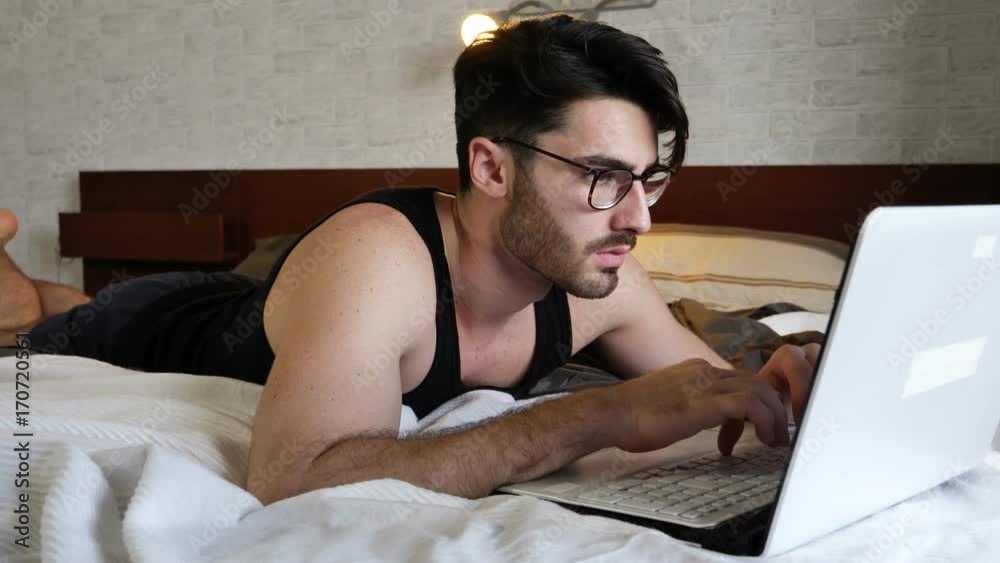 Attractive Young Man with Serious Expression, with Laptop on Bed Working on his Start-up Business - Young Male College or University Student Doing Homework, in Bedroom Attractive Young Man with Serious Expression, with Laptop on Bed Working on his Start-up Business - Young Male College or University Student Doing Homework, in Bedroom