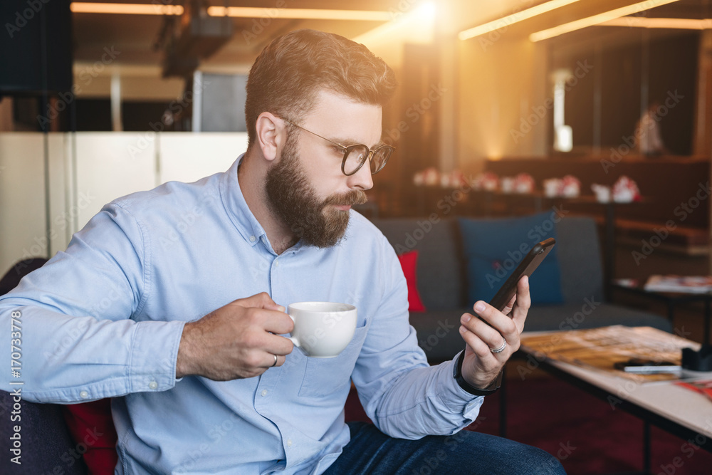 Young serious attractive bearded businessman in blue shirt and glasses sits on sofa in cafe, drinks coffee and uses smartphone