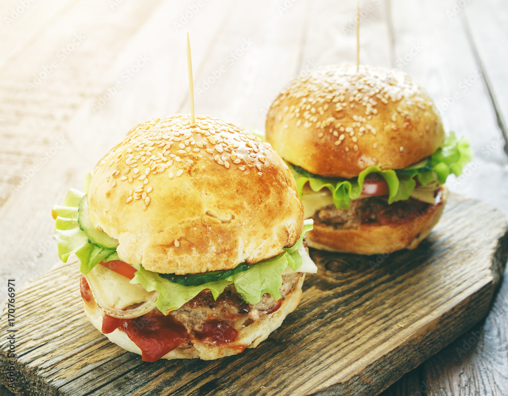 two homemade burgers with green salad leaf, cheese and beef on old rustic wood cutting board in sunlight two homemade burgers with green salad leaf, cheese and beef on old rustic wood cutting board in sunlight