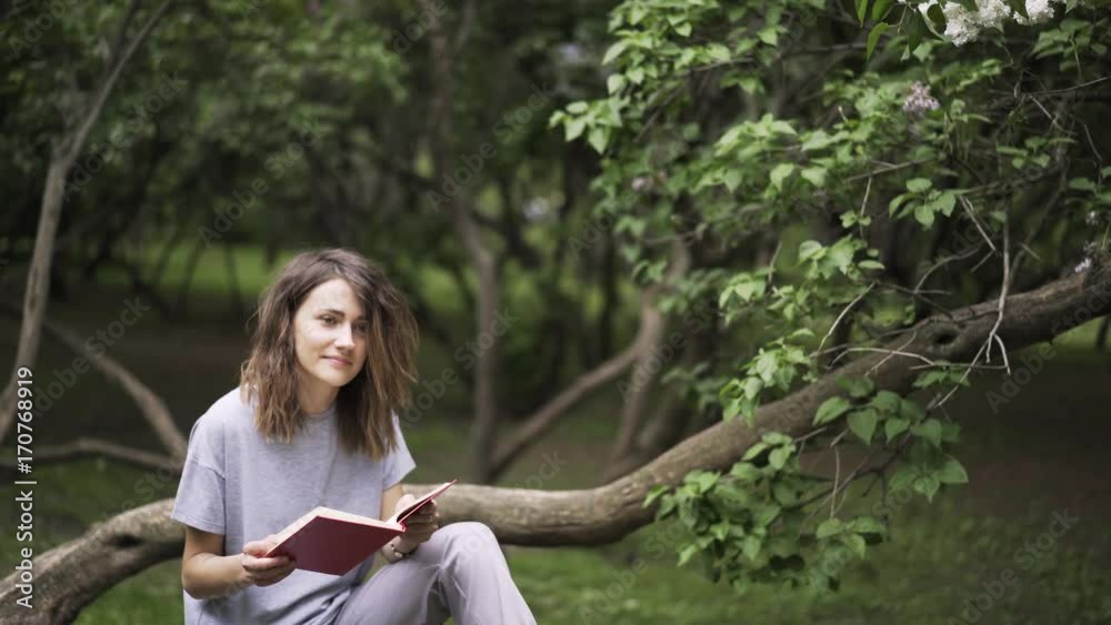 Beautiful young woman reading a red book in a park smiling while sitting on a large tree branch. Tilt down real time establishing shot Beautiful young woman reading a red book in a park smiling while sitting on a large tree branch. Tilt down real time establishing shot