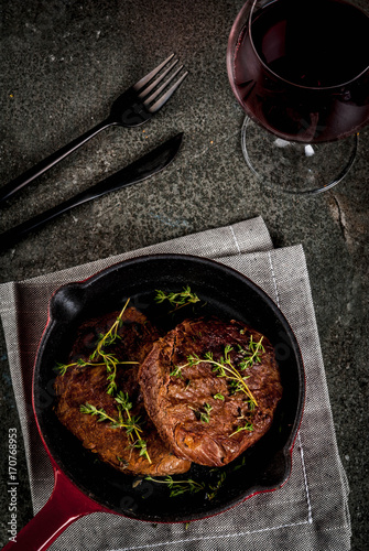 Lunch for one person. Homemade grilled beef steaks with thyme in a portioned frying pan, with a fork, knife and a glass of wine.  On black stone table, copy space top view
