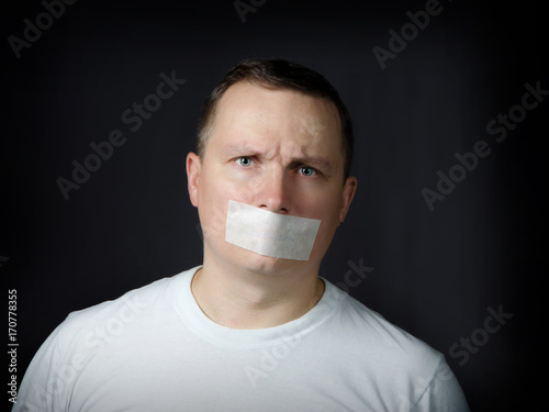 Adult man with a tape pasted over his mouth against a dark background.