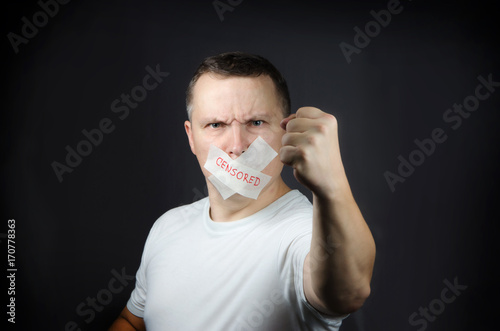 An adult man with a mouth sealed with an adhesive tape, against a dark background. Illustration of censorship.