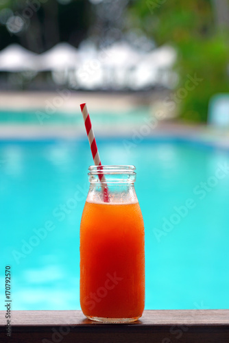 A carrot and orange juice in a glass bottle with a straw by a pool