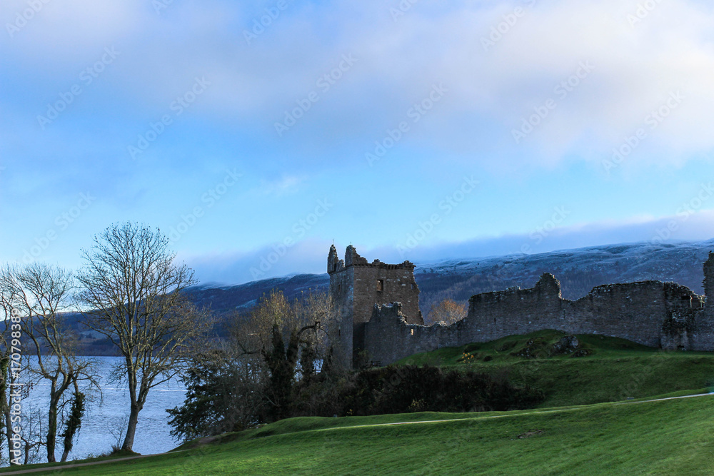 Snowy winter's view of Urquhart Castle with barren trees and Loch Ness in the background.