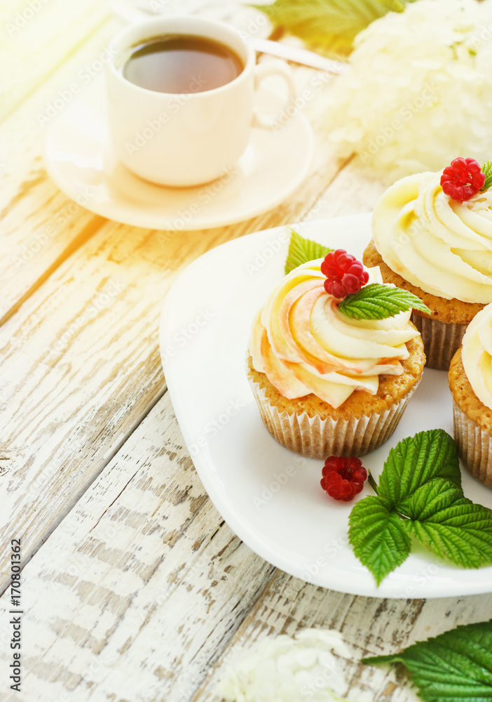 Delicate vanilla cupcakes with cream and raspberries on a white wooden background with a cup of coffee Delicate vanilla cupcakes with cream and raspberries on a white wooden background with a cup of coffee