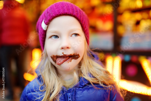 Adorable little girl eating waffle covered with chocolate icing on traditional Christmas market