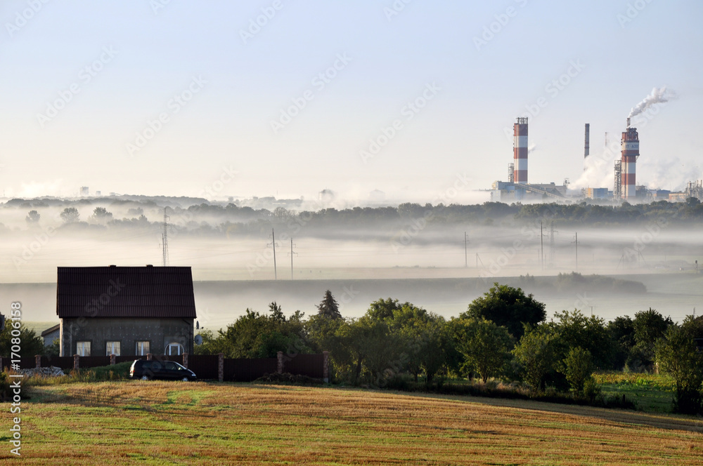 Pipes of working Nitrogen plant in smoke in summer morning.  Abandoned village in the foreground. Grodno, Belarus.