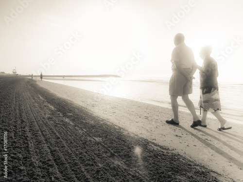 two aged people walking next to the other long the shoreline of an Italian beach in the early morning