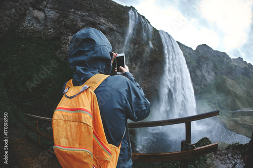 Girl in waterproof clothing stands under the Seljalandsfoss waterfall in Iceland. back view, woman with small orange backpack take a photo on smartphone