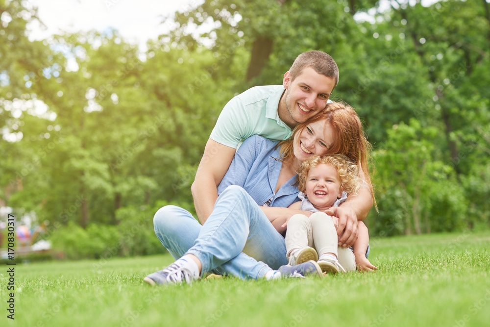 Happy young man smiling joyfully embracing his beautiful wife and daughter sitting on the grass together copyspace family love emotions weekend enjoyment affection parents marriage.