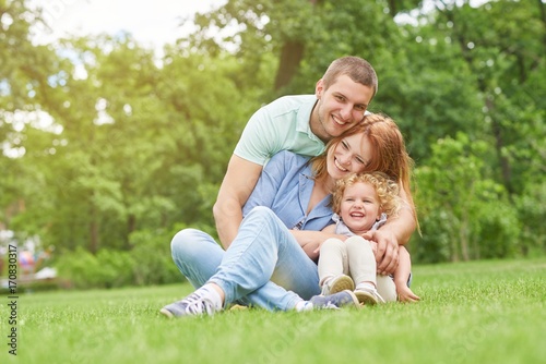 Happy young man smiling joyfully embracing his beautiful wife and daughter sitting on the grass together copyspace family love emotions weekend enjoyment affection parents marriage. © serhiibobyk