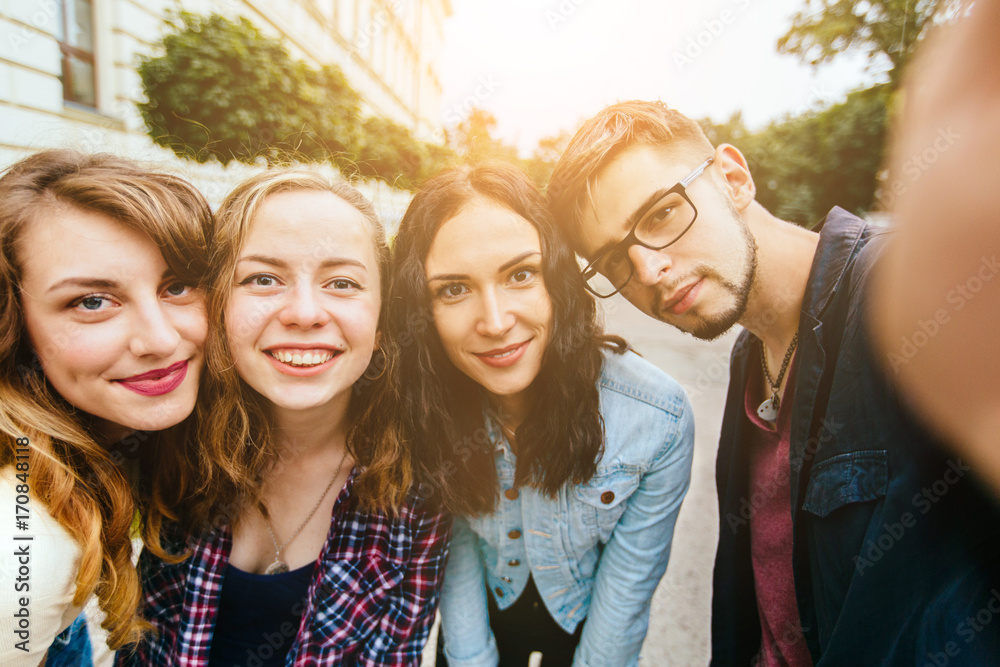 Obraz premium Group of teenager students doing selfie over the university building huggings, smiling, back to school concept. Sun glare effect. Four young happy student making selfie and smiling outdoors.