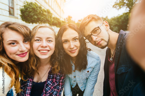 Group of teenager students doing selfie over the university building huggings, smiling, back to school concept. Sun glare effect. Four young happy student making selfie and smiling outdoors.
