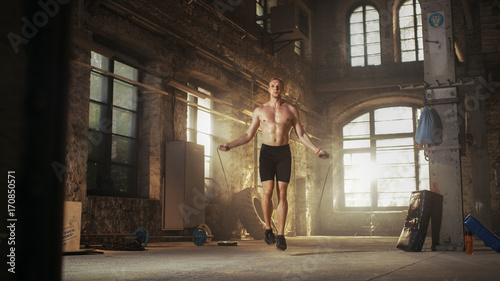 Athletic Shirtless Fit Man Exercises with Jump / Skipping Rope in a Deserted Factory Hardcore Gym. He's Covered in Sweat from His Intense Cross Fitness Training. © Gorodenkoff