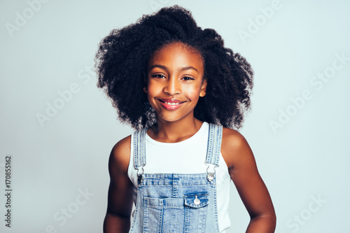 Smiling young African girl wearing dungarees against a gray back