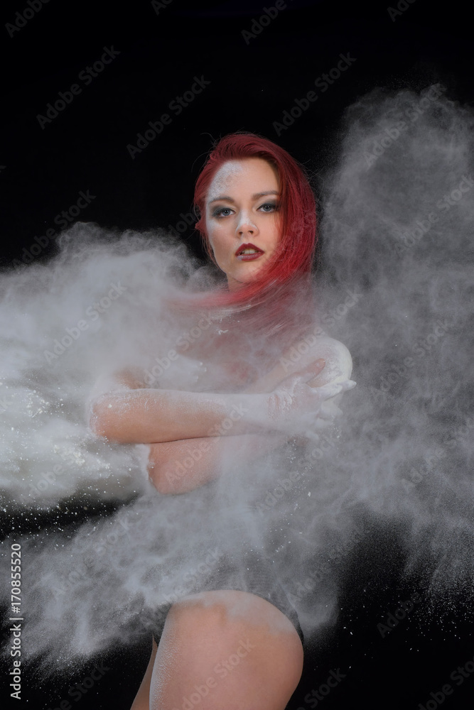 A beautiful redheaded young girl poses  in a studio in front of a black background. As she poses large amounts of white flour  fall on her body as if it were snowfall.    