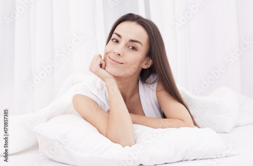 Young beautiful brunette woman lying on pillow in white bed and waking up.