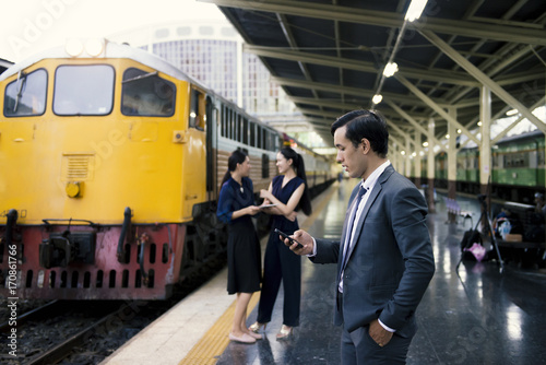 Business man stand at railway playing mobile with couple women and yellow train in background.