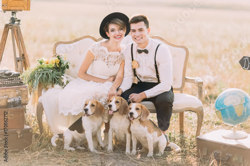 Happy newlyweds are sitting with the three little dogs on the old-fashioned sofa surrouned by the vintage suitcases, flowers and globe. The sunny field composition. photo