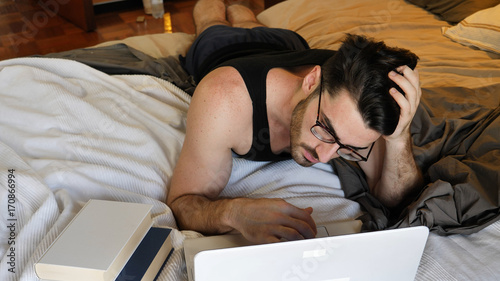 Attractive Young Man with Serious Expression, with Laptop on Bed Working on his Start-up Business, with Books Next to Him - Young Male College or University Student Doing Homework, in Bedroom