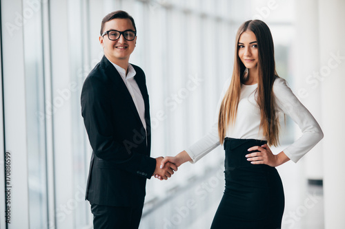 Businessman and businesswoman shaking hands together while standing in front of office building windows