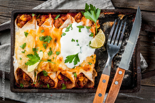 Mexican food. Cuisine of South America. Traditional dish of spicy beef enchiladas with corn, beans, tomato. On a baking tray, on old rustic wooden background. Top view photo