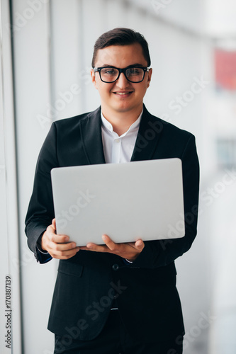 Young smiling businessman using laptop on corporate location, indoor neat panoramic window