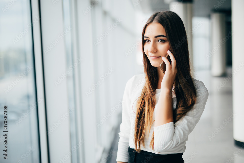 Happy businesswoman standing in her office in a highrise building overlooking the cityscape. Businesswoman talking on mobile phone standing near window in office. Happy businesswoman standing in her office in a highrise building overlooking the cityscape. Businesswoman talking on mobile phone standing near window in office.