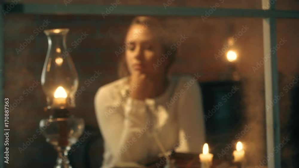Rack focus scene through a window to a young pensive pioneer woman lit by the light from candles and oil lamps