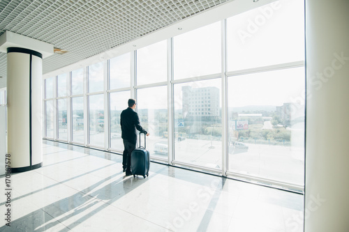 Businessman with suitcase at airport international departure gate looking at flying airplane through the windows